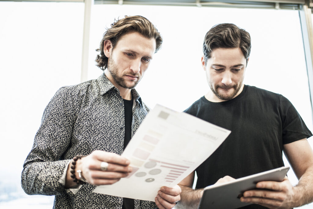 Dos hombres parados en una oficina, mirando una página impresa y refiriéndose a una tableta digital.