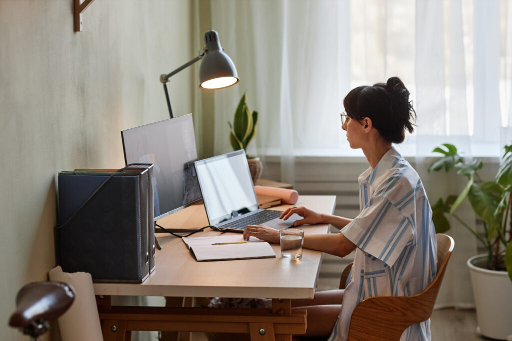 Foto de uma mulher com o cabelo rpeso e uma camisa branca com listras, sentada em frente ao computador em um escritório. 
